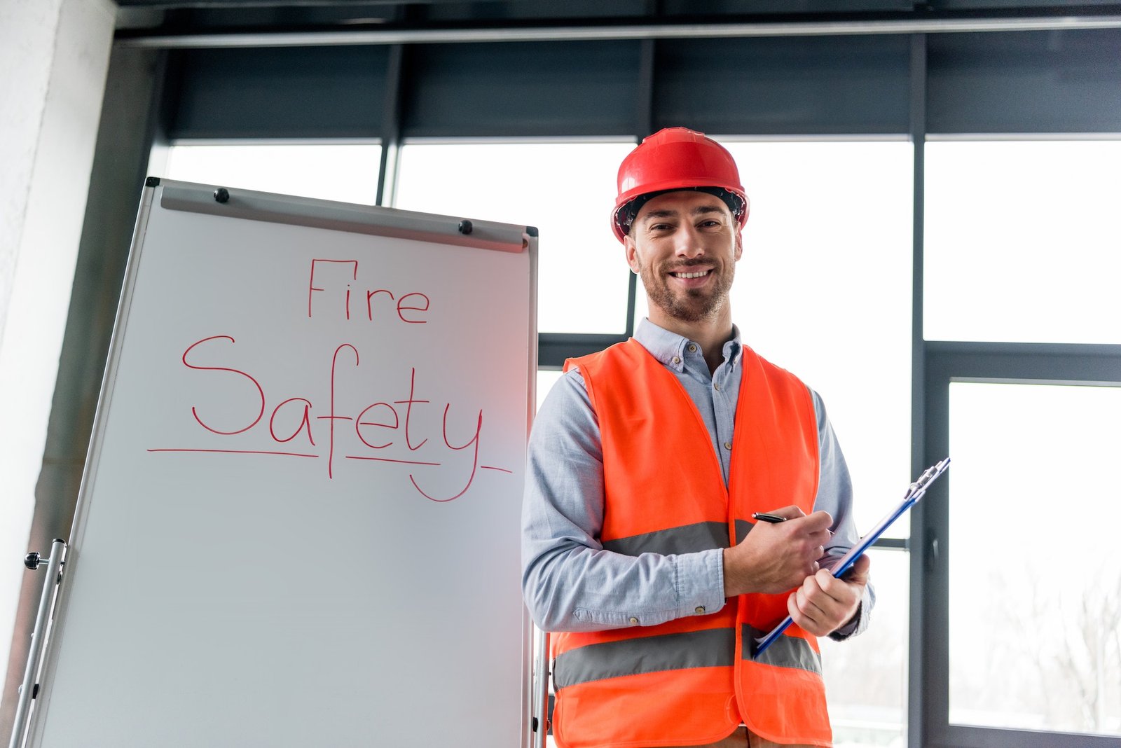 cheerful firefighter in helmet holding clipboard and pen while standing near white board with fire